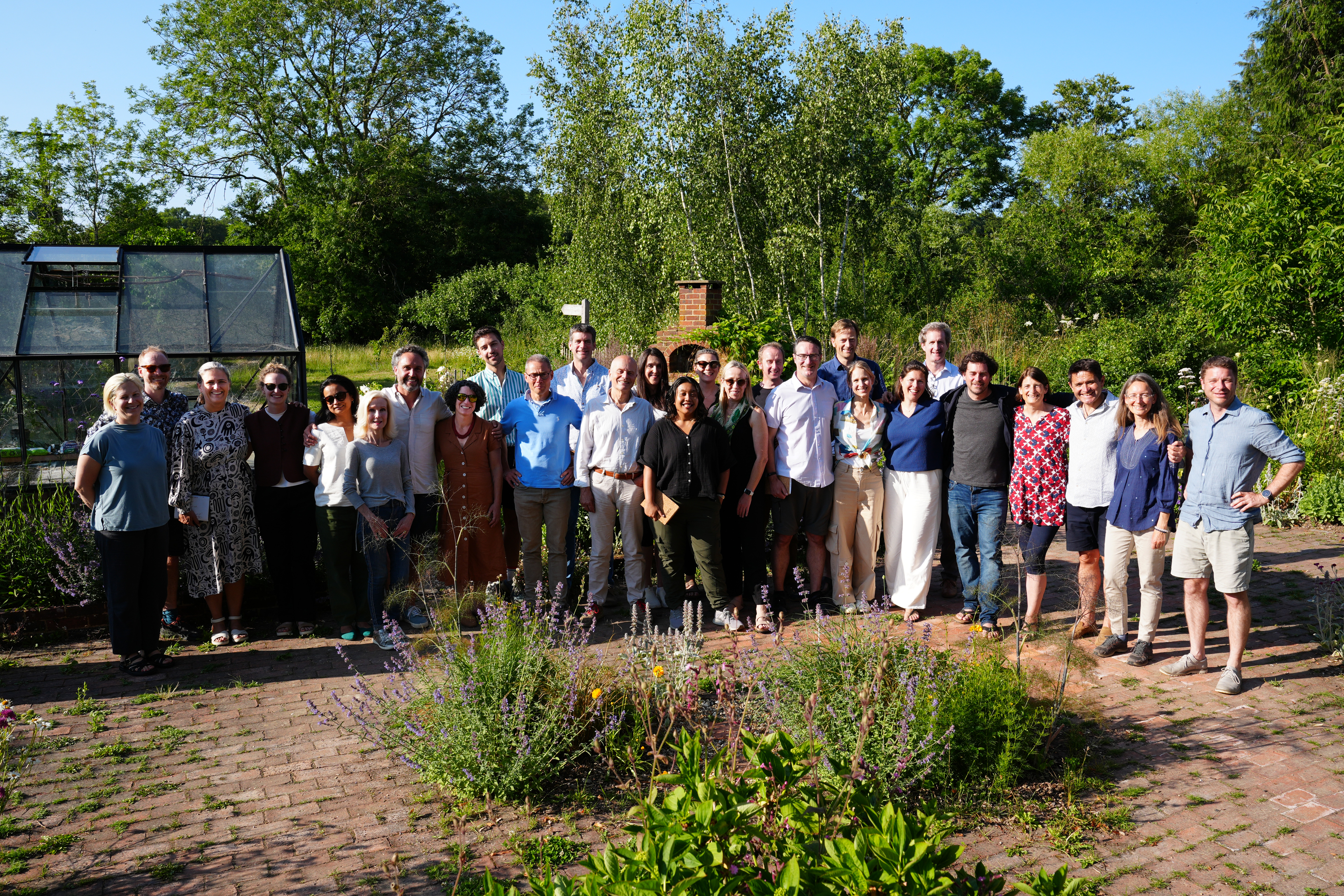 A group photo of the participants of Ostara's Imaginal Studio, standing in a farmyard in front of a greenhouse in the sun.