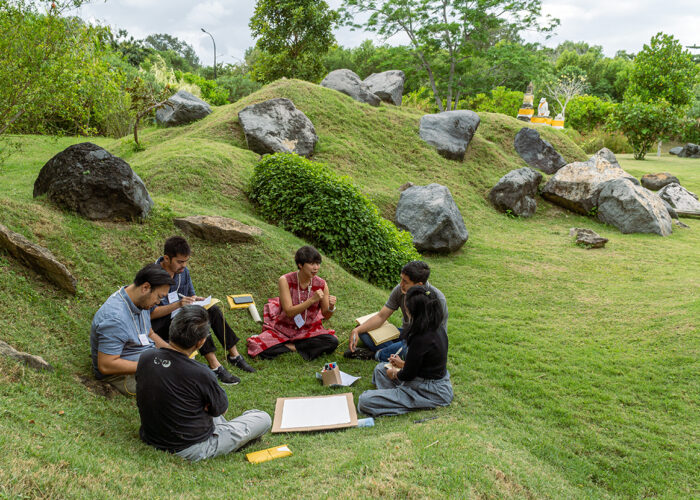 Five People Sit In A Small Circle In A Grassy Landscape, Gathered Around Some Papers.
