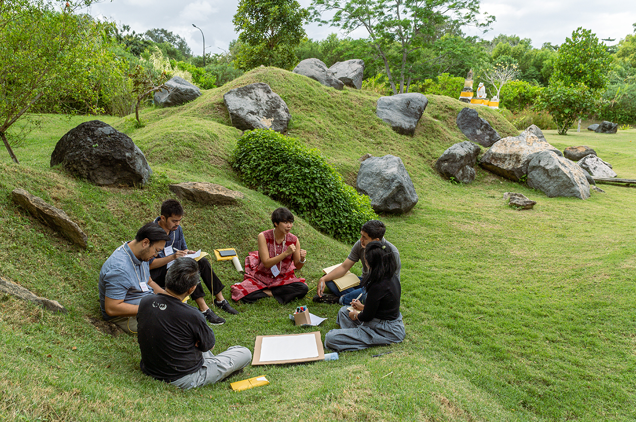 Six people sit in a small circle in a grassy landscape, gathered around some papers. 