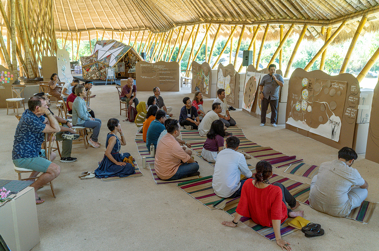 A large group of people sit on mats on a floor under a bamboo roof structure, looking at a presenter at the front.