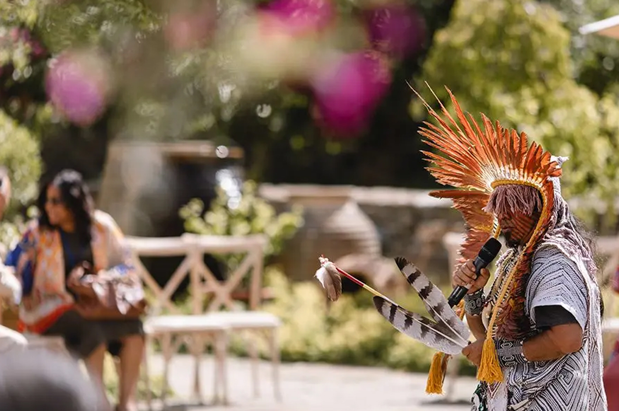 An indigenous leader wearing a large feathered headdress speaks through a microphone.