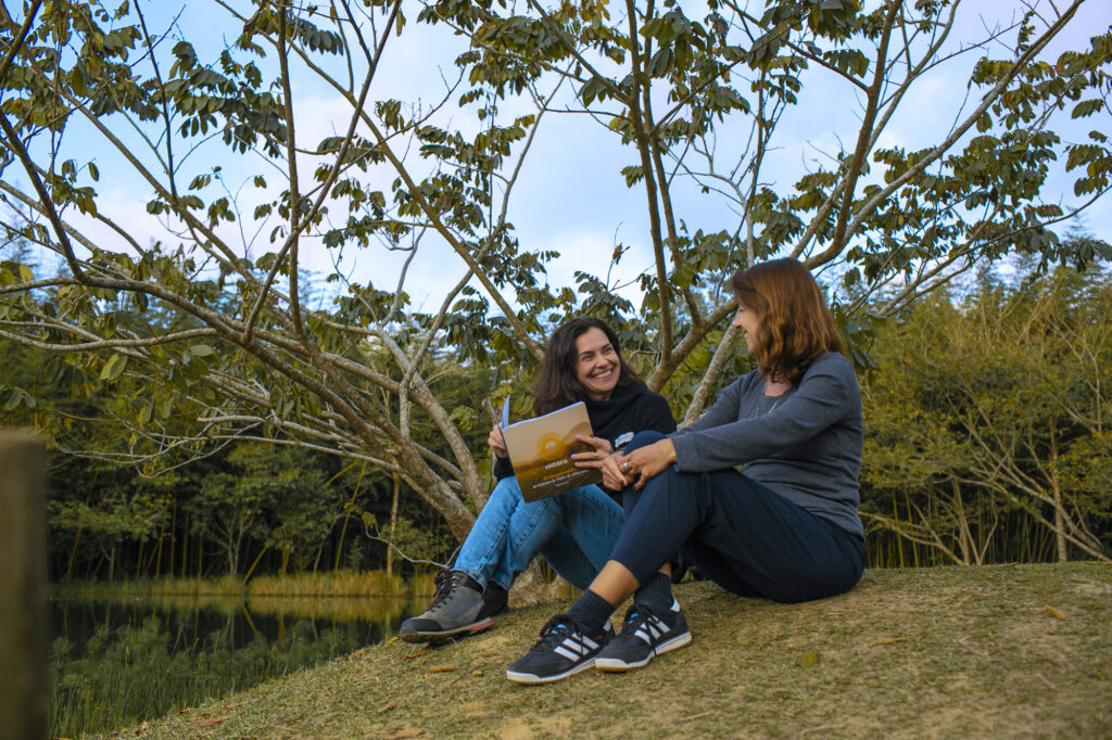 Two white women sit on a grassy verge with trees in the background, deep in discussion. 