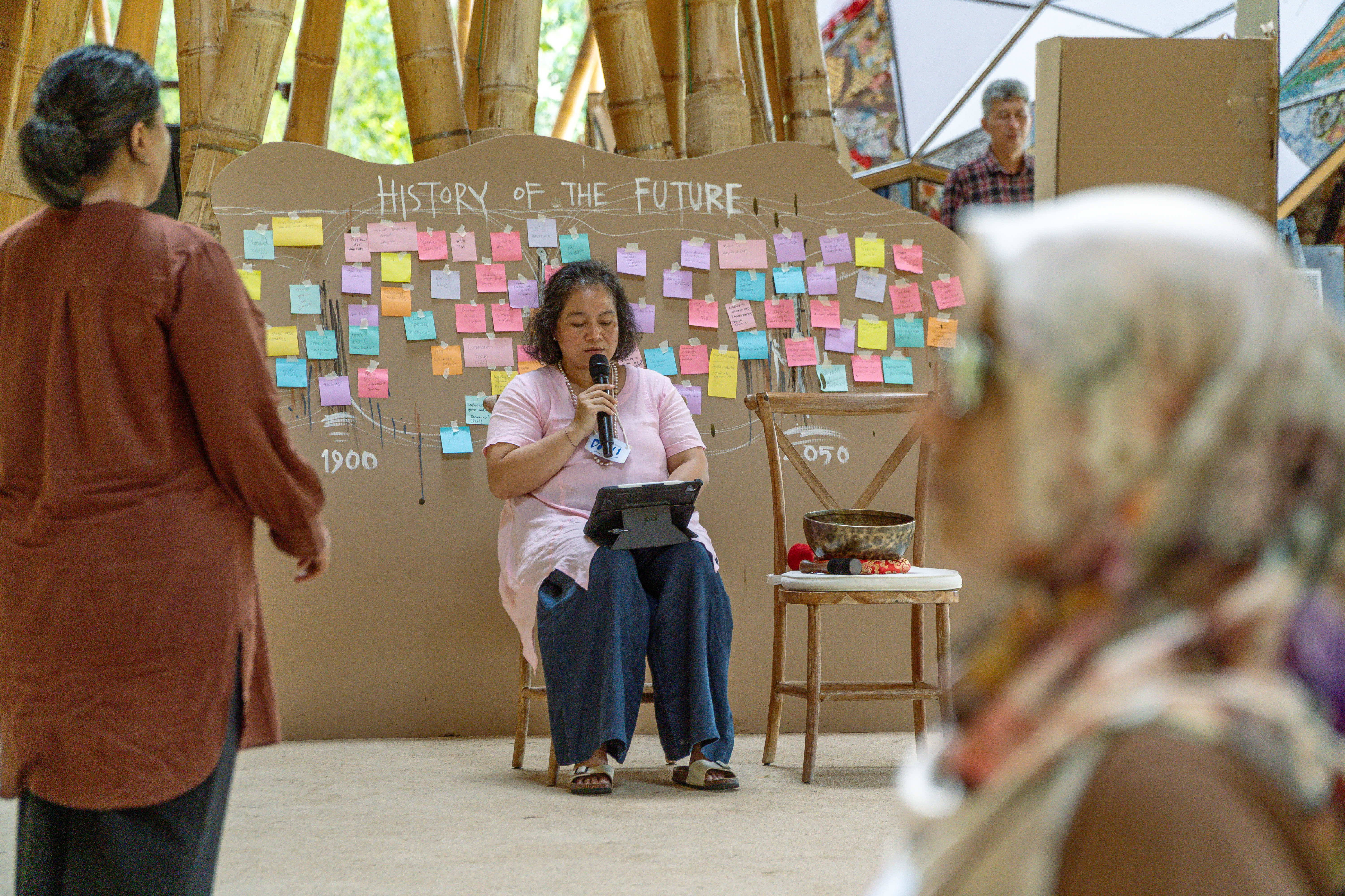 A woman sits on a wooden chair holding a microphone, in front of a large piece of cardboard with the title 'history of the future' written on it, in additional to many coloured post-it notes.