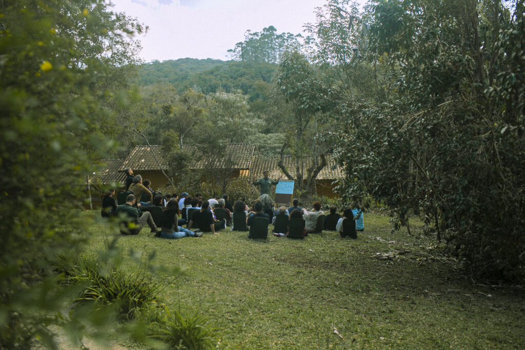 A group of people sit on the grass surrounded by trees, at Ostara's Imaginal Studio in Brazil.