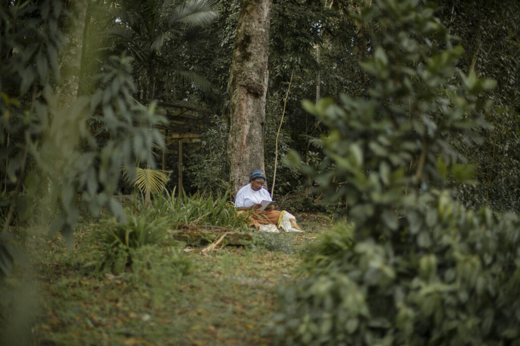 A woman sits under a tree, writing in a notebook, at Ostara's Imaginal Studio in Brazil.